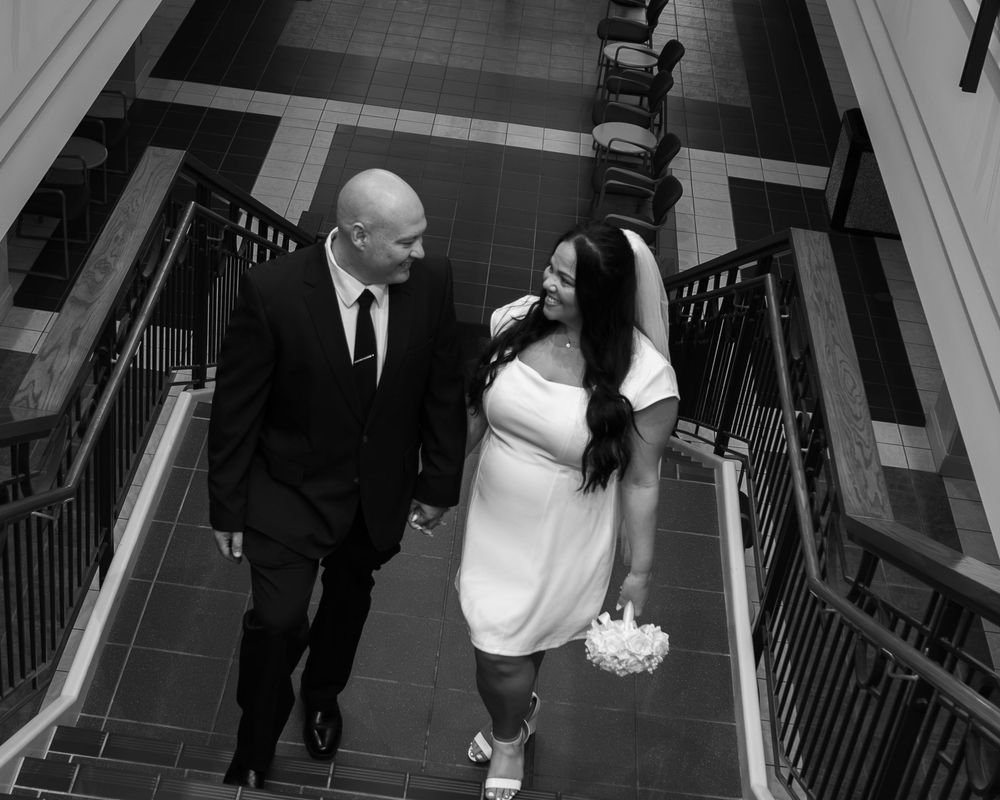 Black and white photo of a bride and groom holding hands on stairs, smiling at each other on their wedding day.