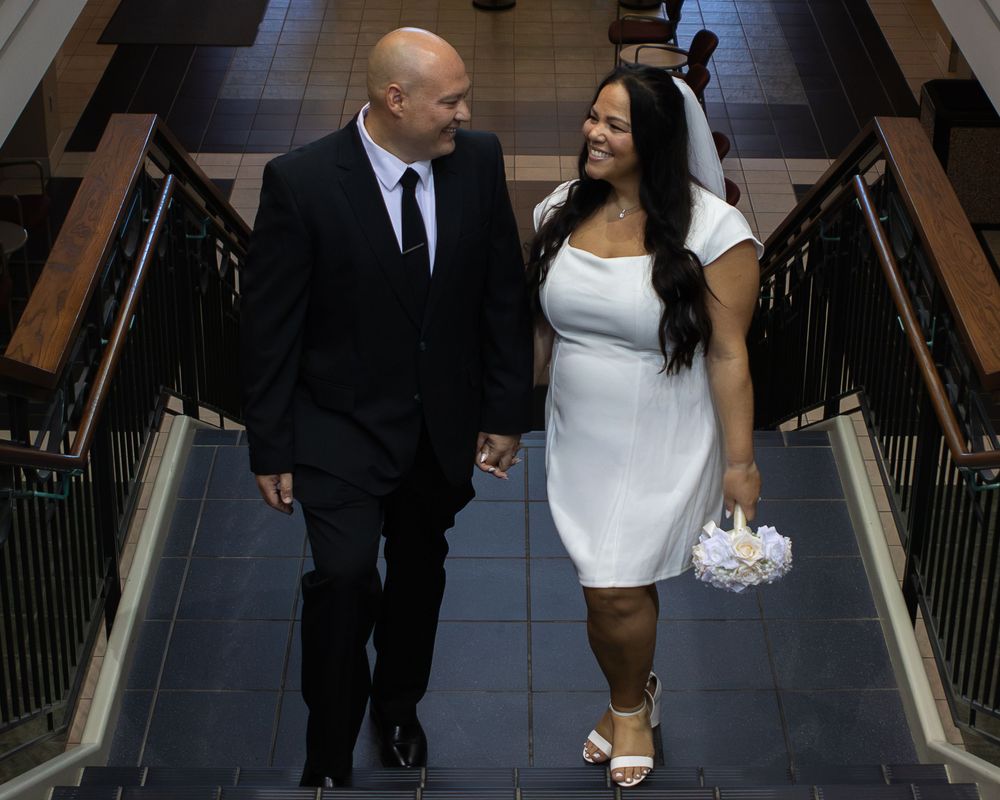 Bride and groom holding hands walking up stairs, bride in white mini dress with veil and bouquet, groom in black suit.