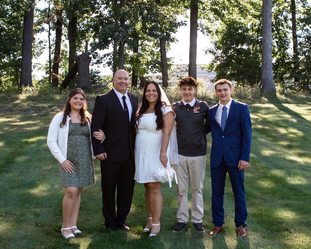 Family of five posing outdoors on green grass with trees, dressed in formal attire including a white wedding dress.