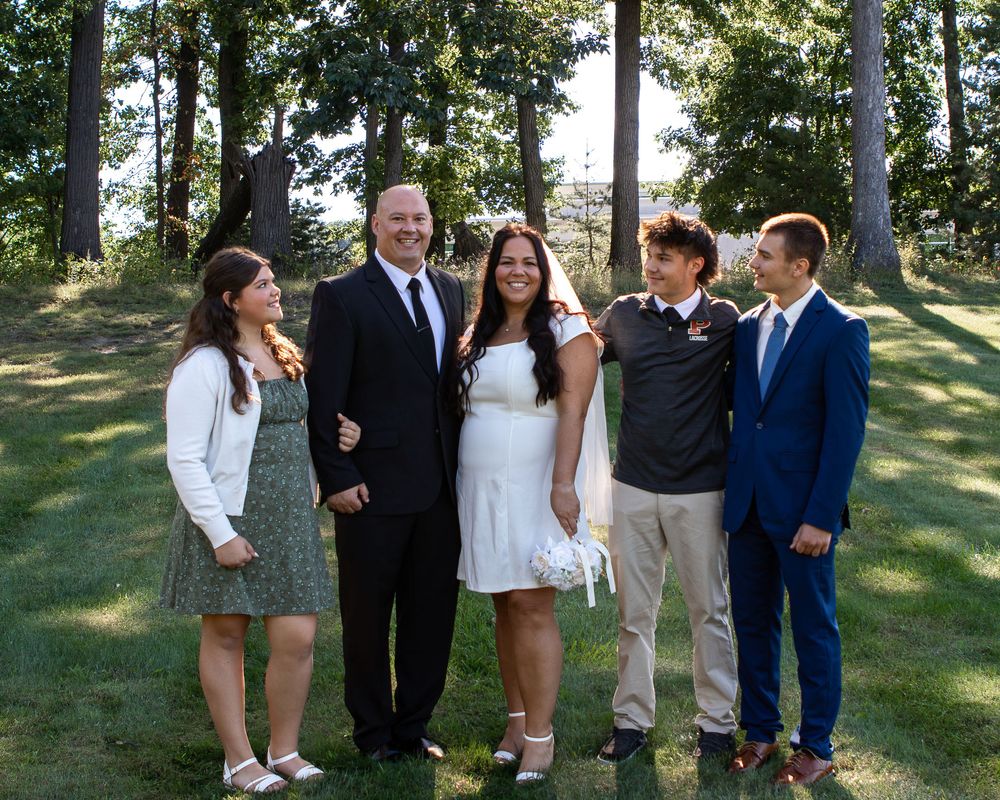 Family of five smiling outdoors in formal attire on a sunny day with green trees in the background.