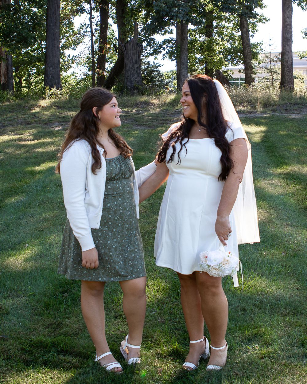 Bride in white mini dress and veil smiles with bridesmaid in green floral dress outdoors.