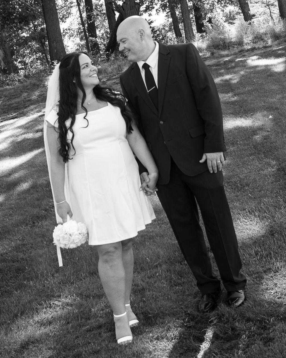 Bride in white dress holding bouquet stands with groom in black suit holding hands outdoors on green lawn.