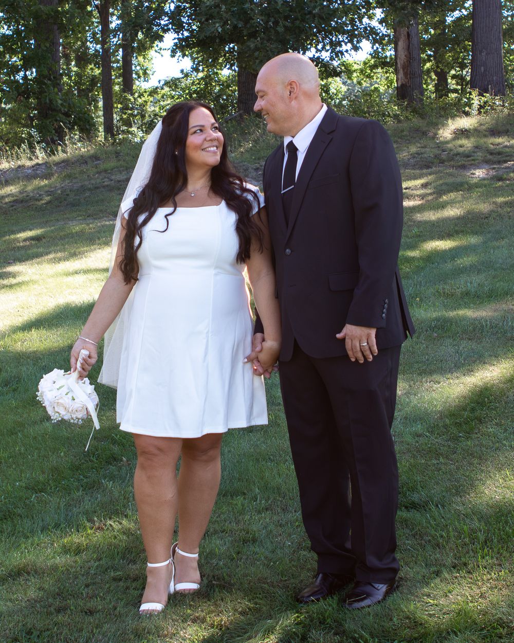 Bride in white dress and groom in black suit holding hands and smiling at each other outdoors on green grass.