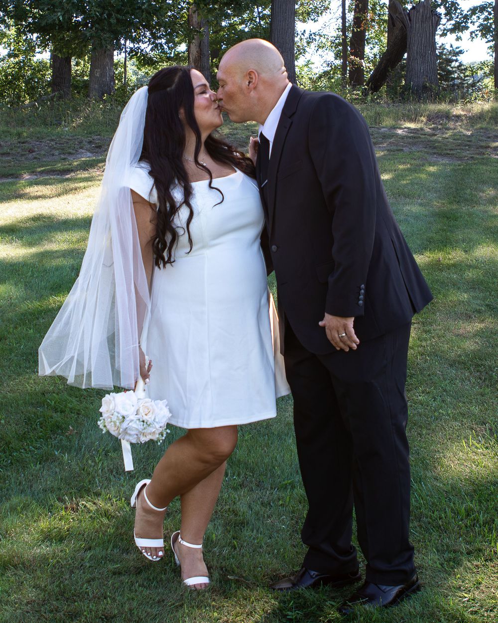Bride and groom share a kiss outdoors, she holds white bouquet wearing veil and white dress.