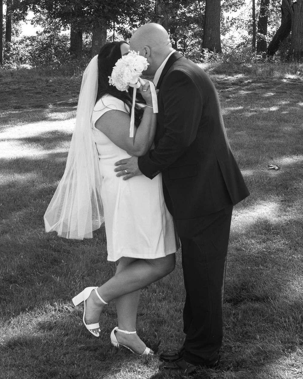 Black and white photo of a bride and groom sharing a kiss outdoors, bride holding white bouquet and wearing veil.