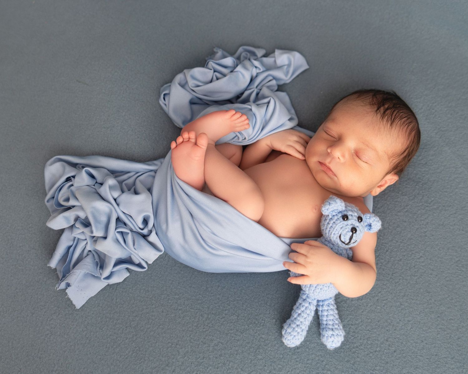 Newborn baby sleeping peacefully wrapped in a blue blanket, holding a small crocheted blue teddy bear on a gray background.