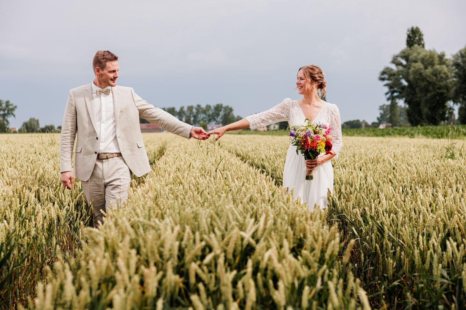 Bride and groom holding hands in a green wheat field, bride holding colorful bouquet, groom in tan suit.
