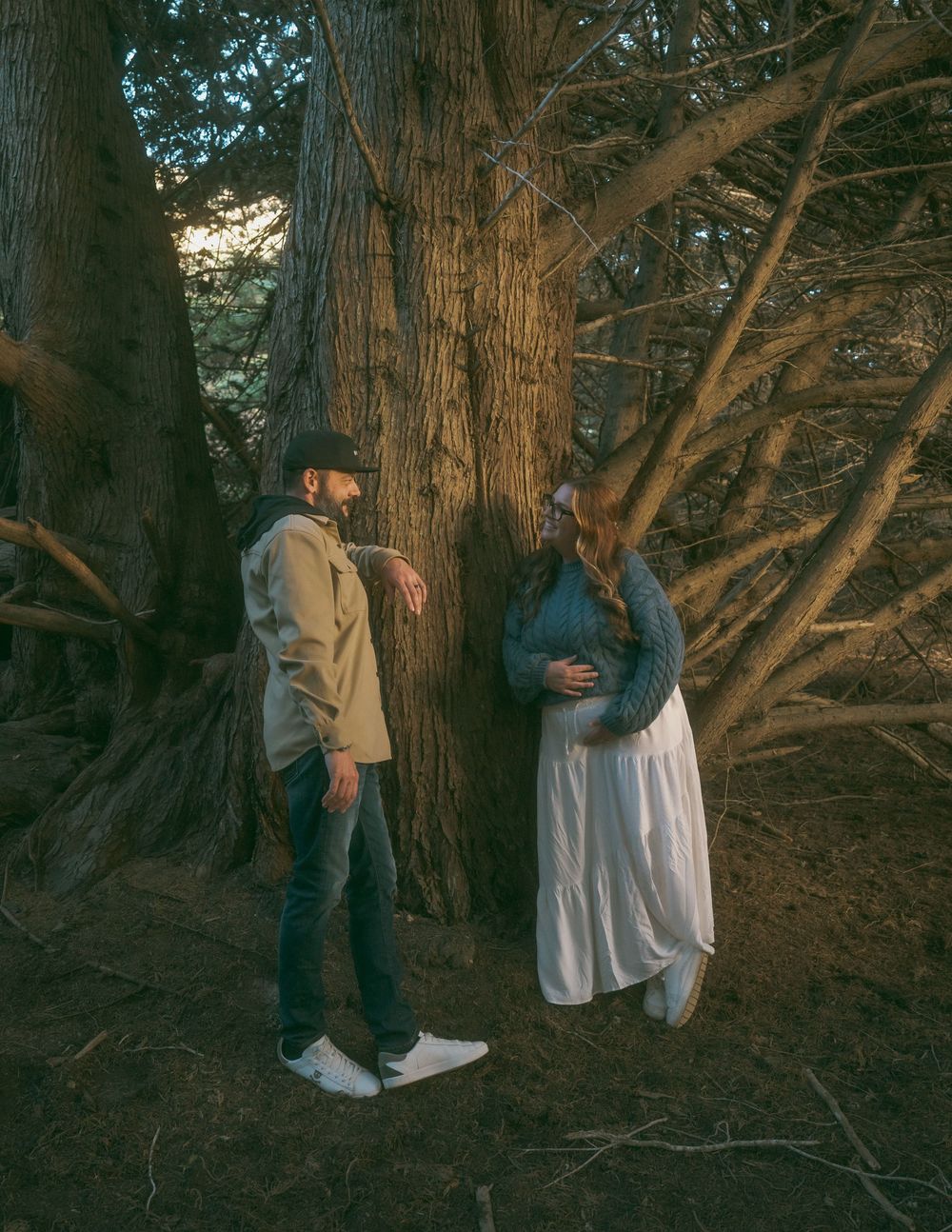 Couple standing by large tree trunk in forest, pregnant woman in white skirt and blue sweater with man in tan jacket.