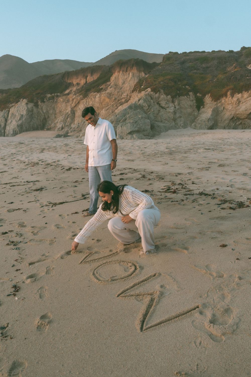 Couple writing 'NOW' in sand on a beach with rocky cliffs, wearing white outfits in golden hour light.
