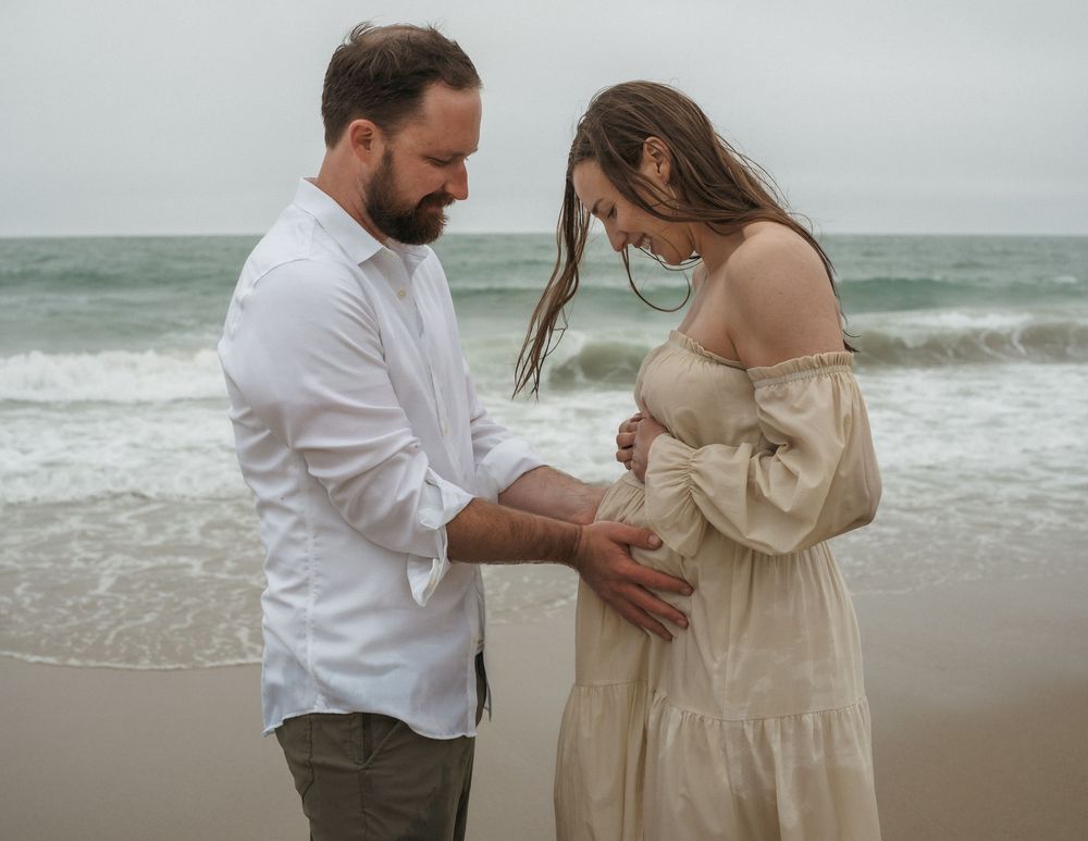 Couple sharing a tender moment on a beach, woman in cream off-shoulder maternity dress, man in white shirt.
