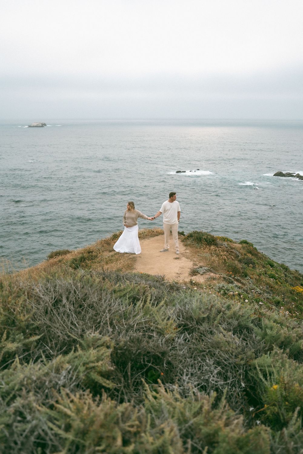 Couple holding hands on a coastal cliff overlooking the ocean on an overcast day.