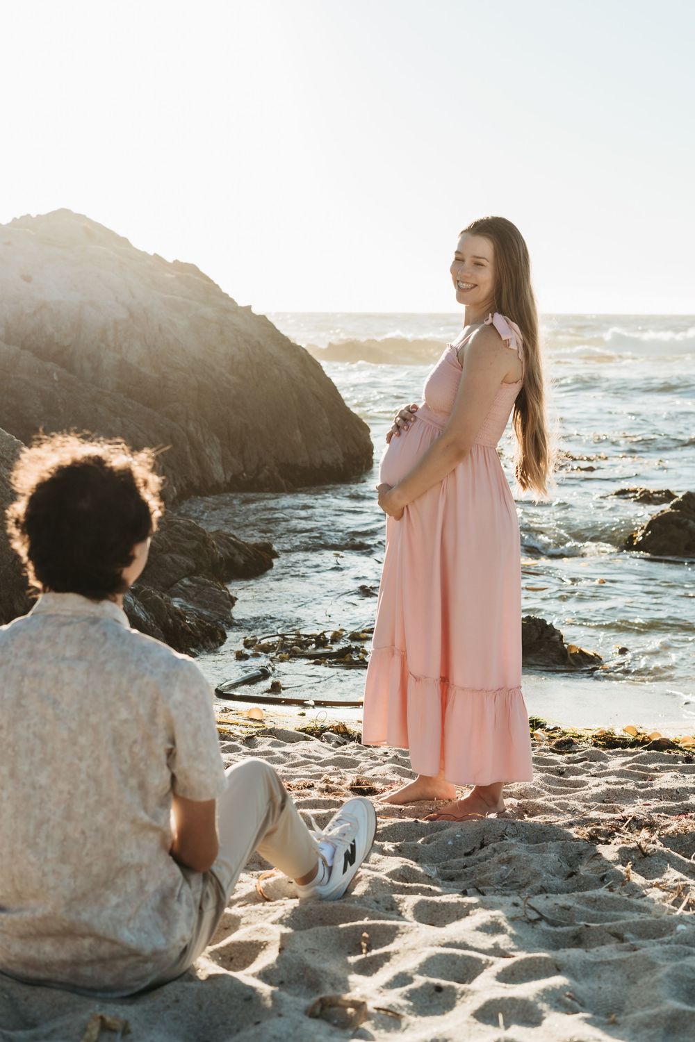 Pregnant woman in pink dress holding belly on sandy beach, partner sitting nearby with ocean rocks backdrop.