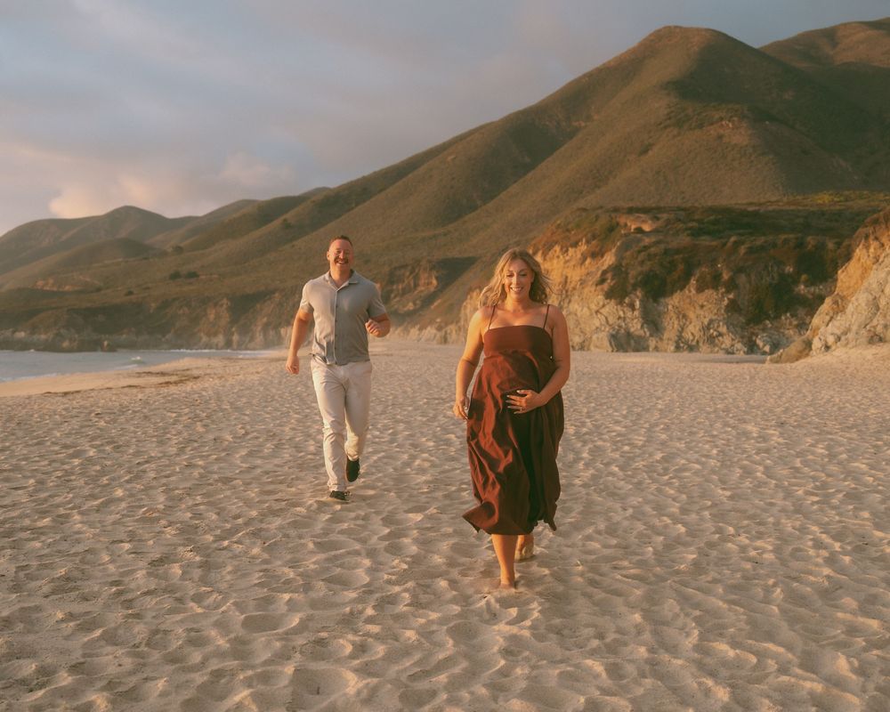 Pregnant woman in brown dress walks on sandy beach with man at golden hour, coastal cliffs in background.