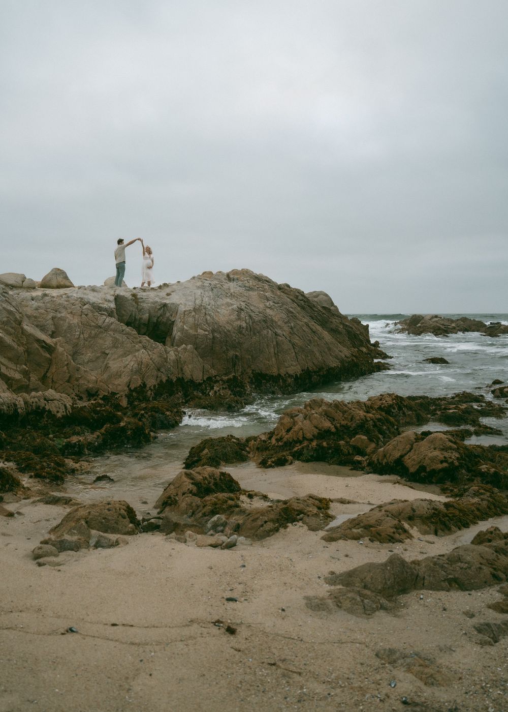 Couple dancing on rocky coastal cliffs by the ocean on an overcast day at a scenic beach.