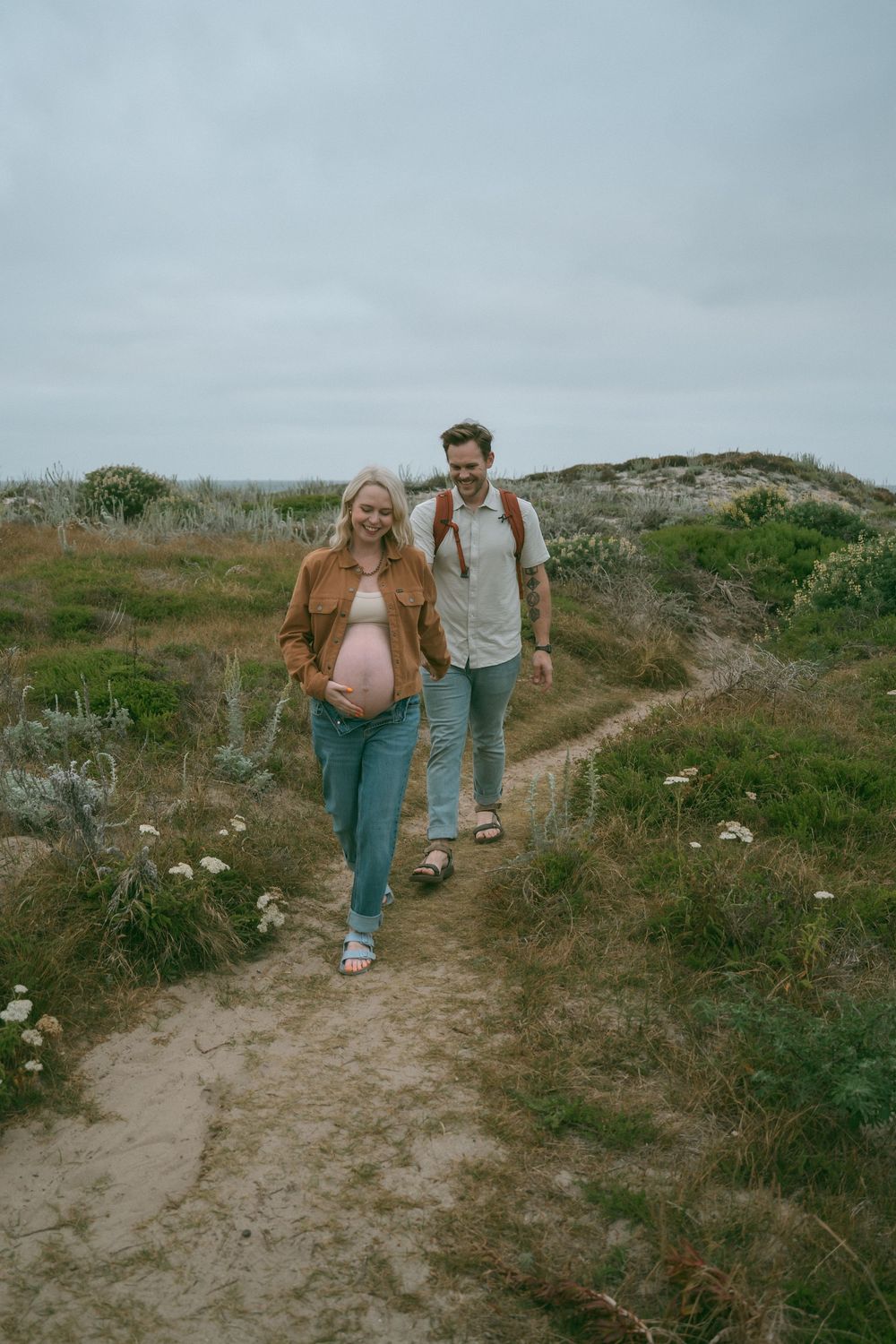A pregnant woman and her partner walk along a scenic coastal trail surrounded by green hills and wildflowers.