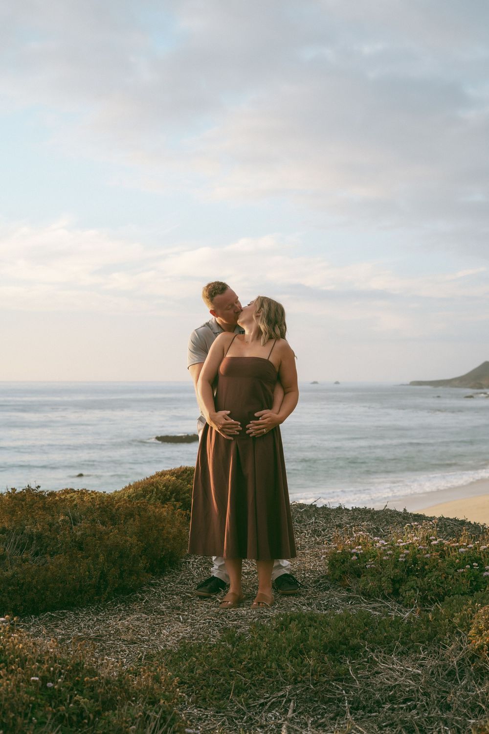 Couple sharing a tender kiss on a coastal bluff, man embracing pregnant woman in brown dress at sunset beach.