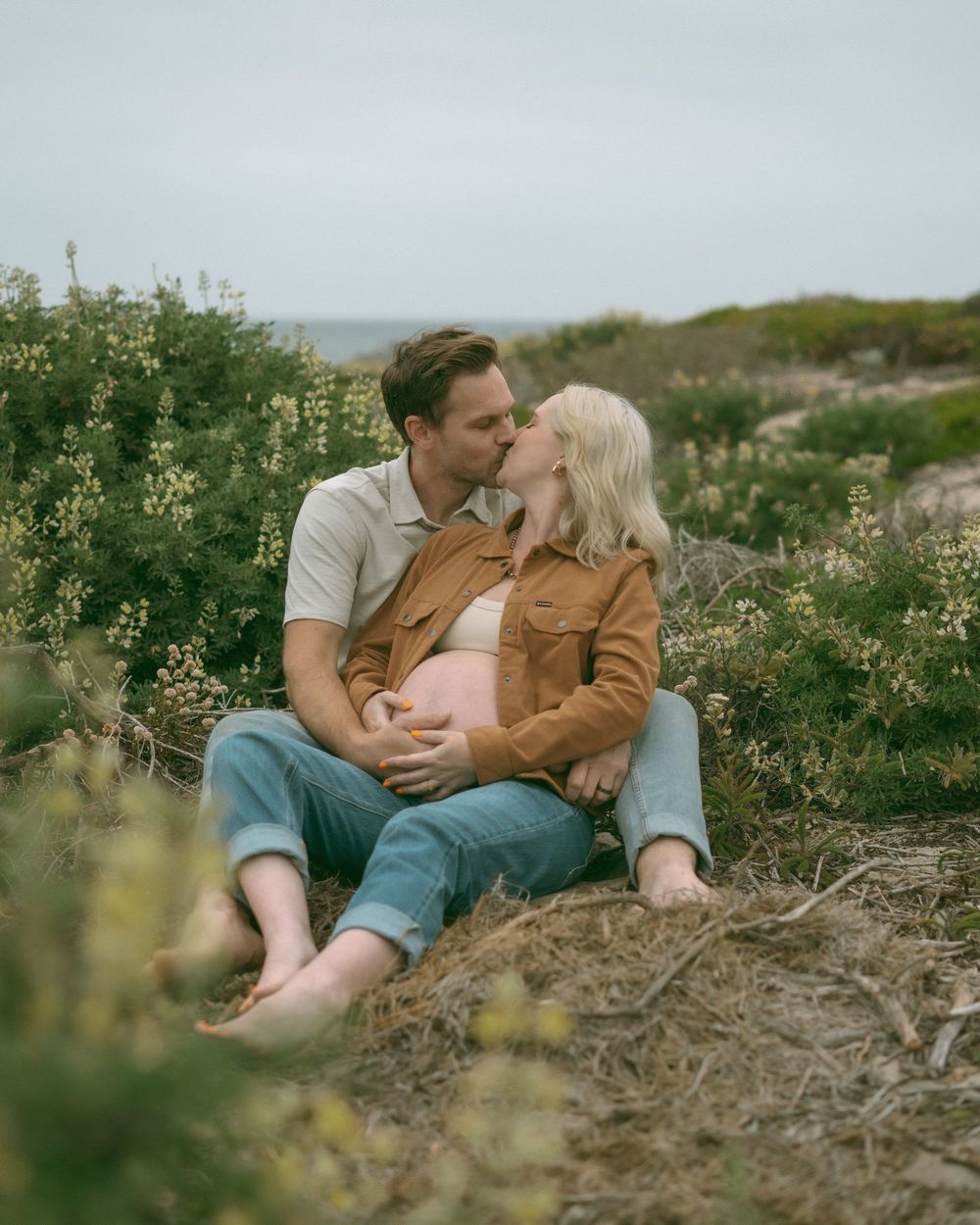 Couple sharing a kiss during an outdoor maternity photo shoot, sitting barefoot in coastal shrubs.