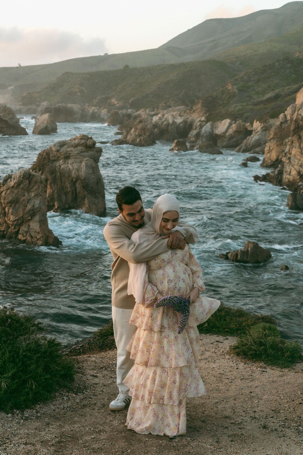Couple embracing on rocky Big Sur coastline, woman in floral hijab dress, man in beige suit, ocean waves behind them.
