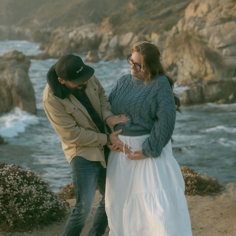 Couple sharing a tender moment on a rocky coastal cliffside, woman in white skirt and blue sweater, man in hat and jacket.