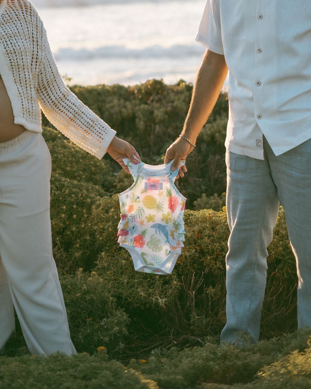 Couple holding a floral baby onesie together outdoors near the ocean, announcing their pregnancy.