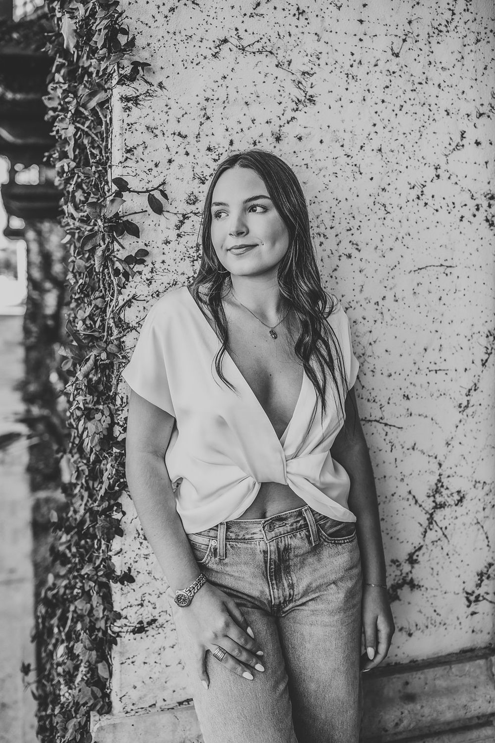 Black and white portrait of a young woman in jeans and white top leaning against a textured wall with ivy.
