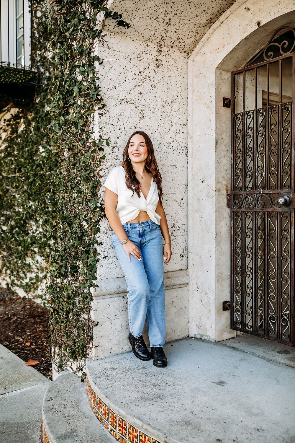 Woman in white top and jeans leaning against a textured wall near an ornate iron gate.
