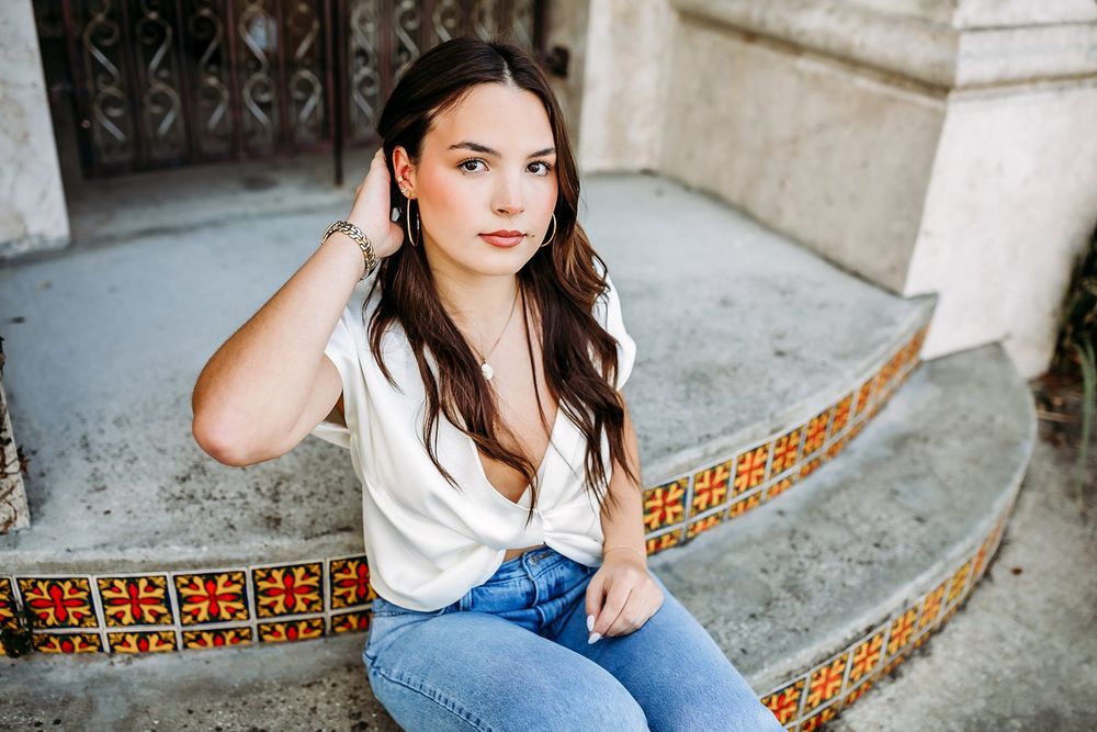 Young woman in white top and jeans sitting on decorative tiled steps outdoors, posing for a portrait photo.