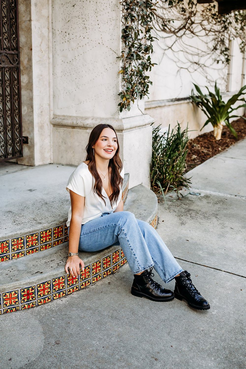Young woman smiling, sitting on decorative tiled steps in jeans, white tee, and black boots near a building.