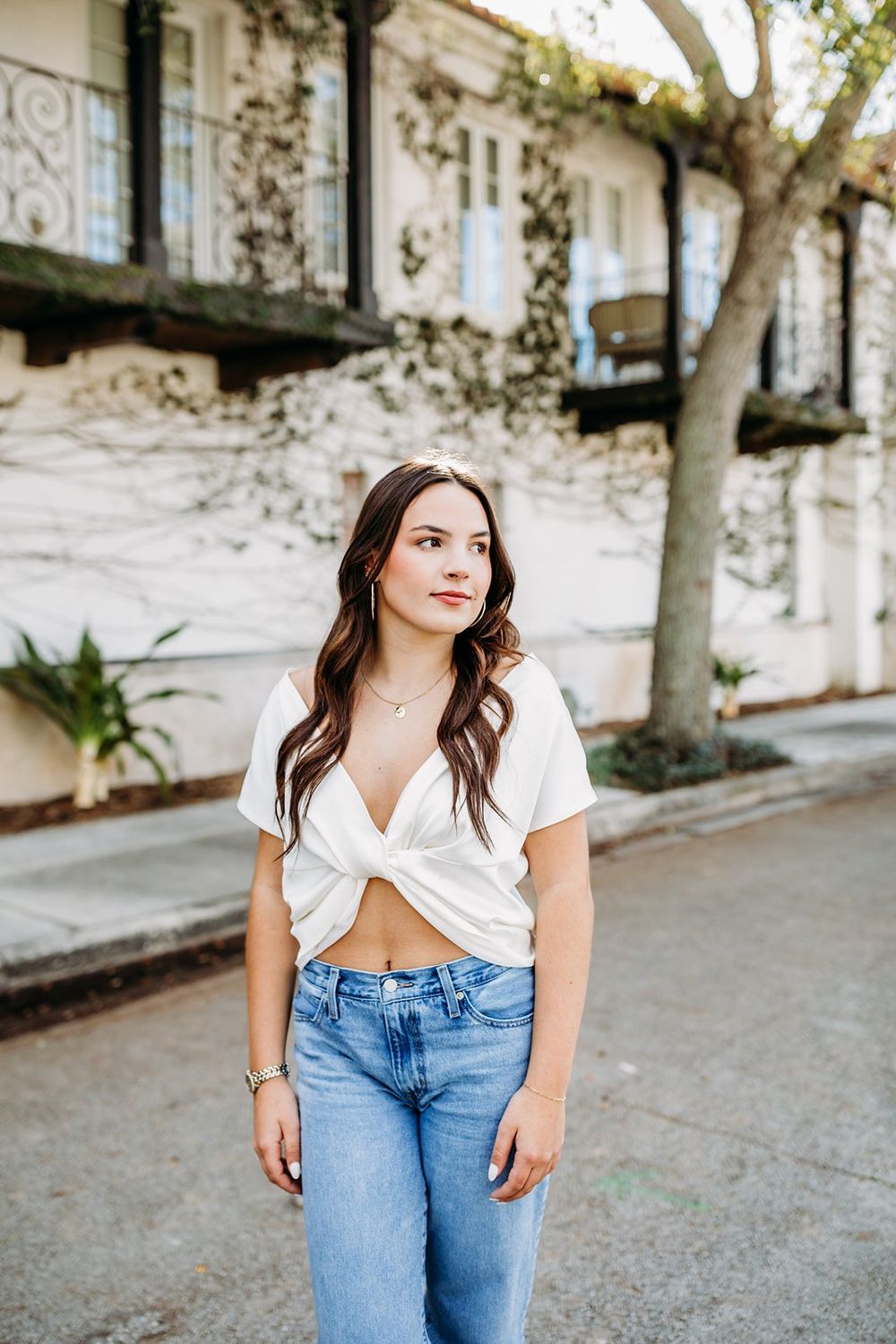 Young woman in white crop top and jeans posing outdoors near a white building with balconies and trees.
