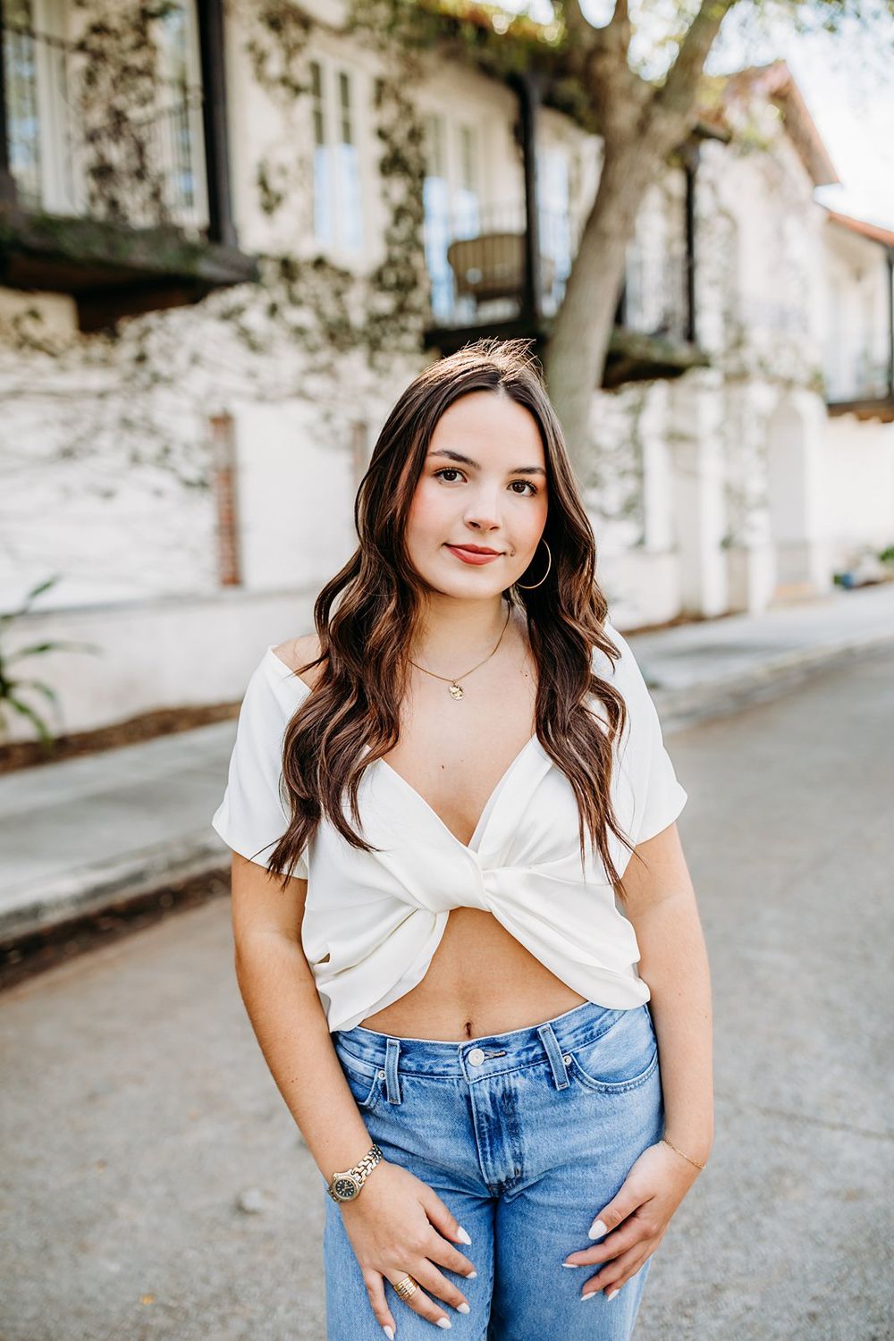 Young woman in white crop top and jeans posing on a charming street with ivy-covered buildings.