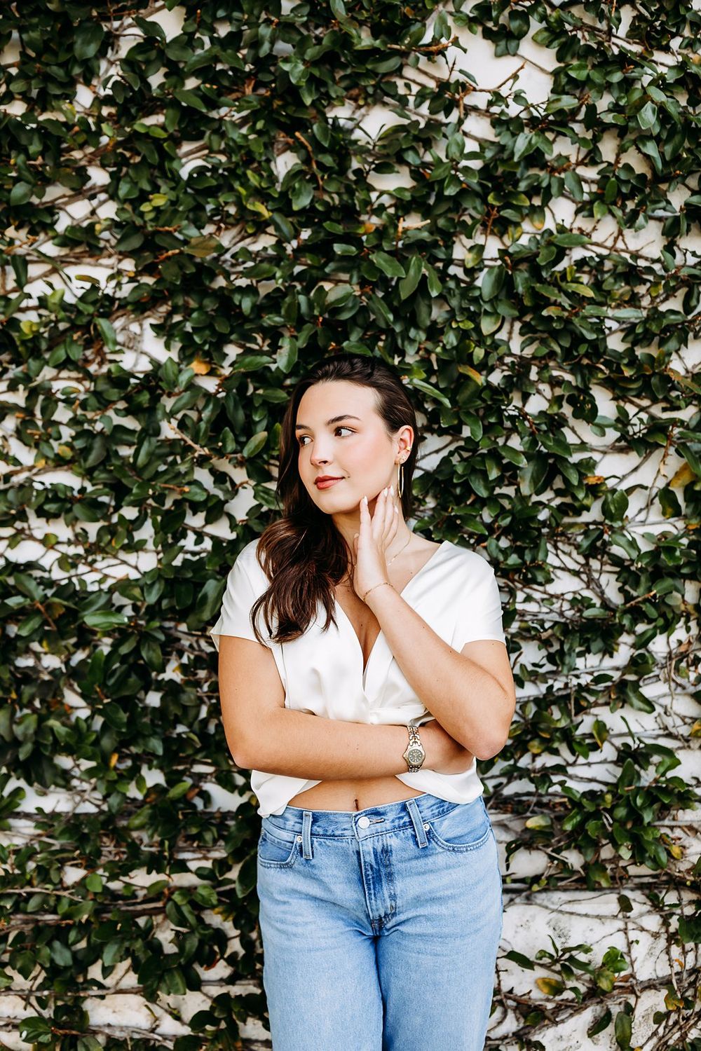 Young woman in white top and jeans poses thoughtfully against a lush green ivy-covered wall.