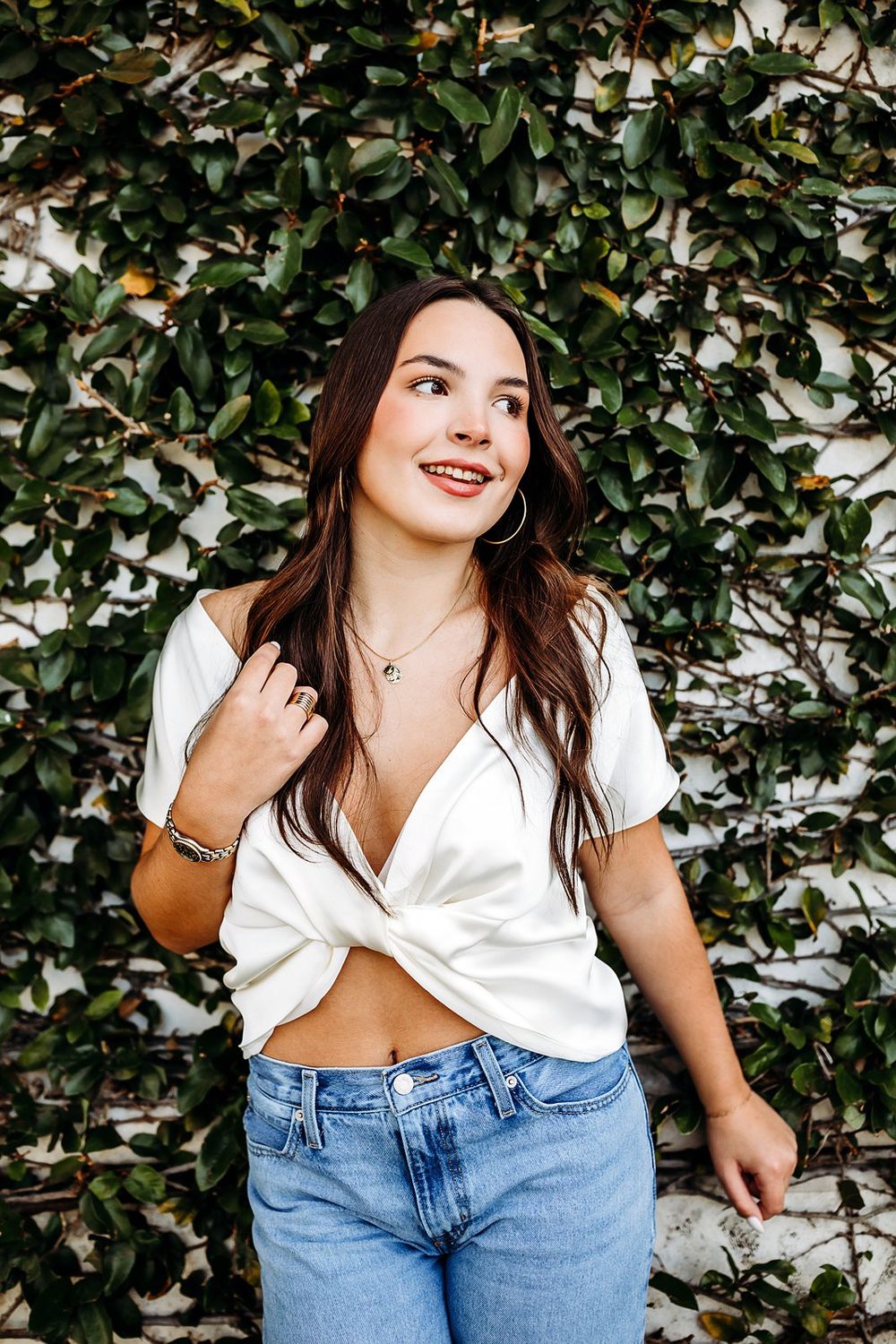 Young woman in white crop top and jeans smiling outdoors against a lush green ivy-covered wall.