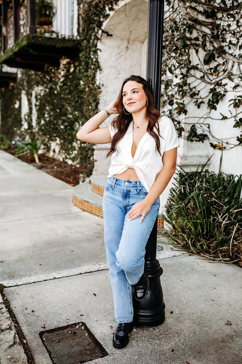 Young woman in white crop top and light blue jeans leaning on black post outdoors near ivy-covered building.