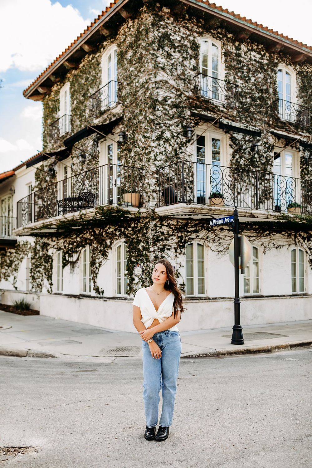 Young woman in white top and jeans poses on a street corner before an ivy-covered historic building with iron balconies.