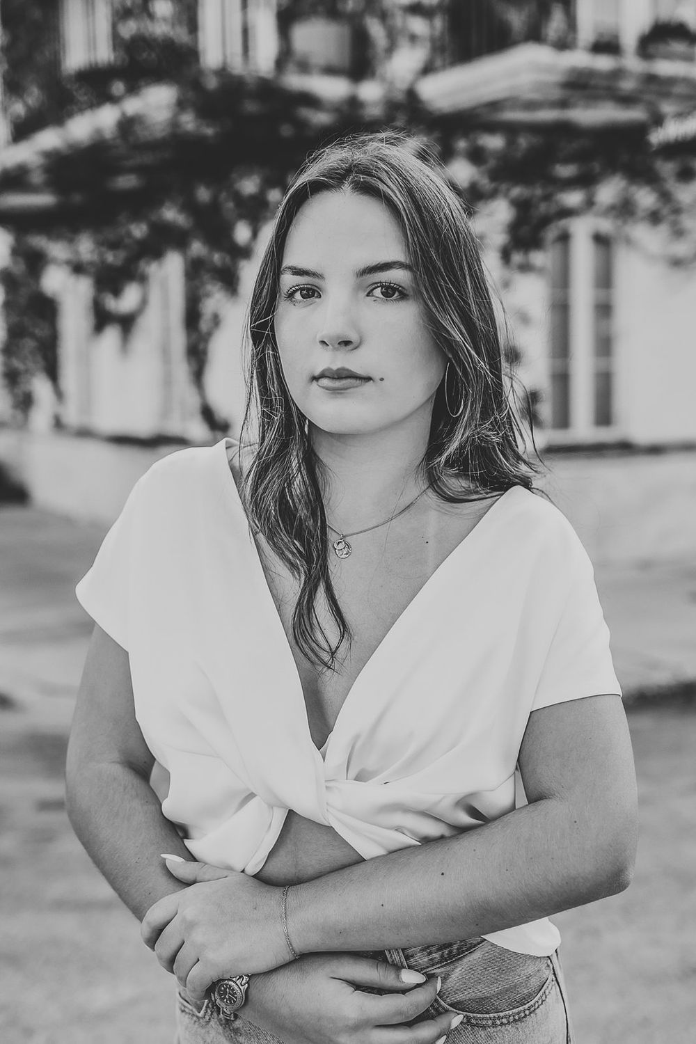 Black and white portrait of a young woman in a white top, arms crossed, standing outdoors near a building.