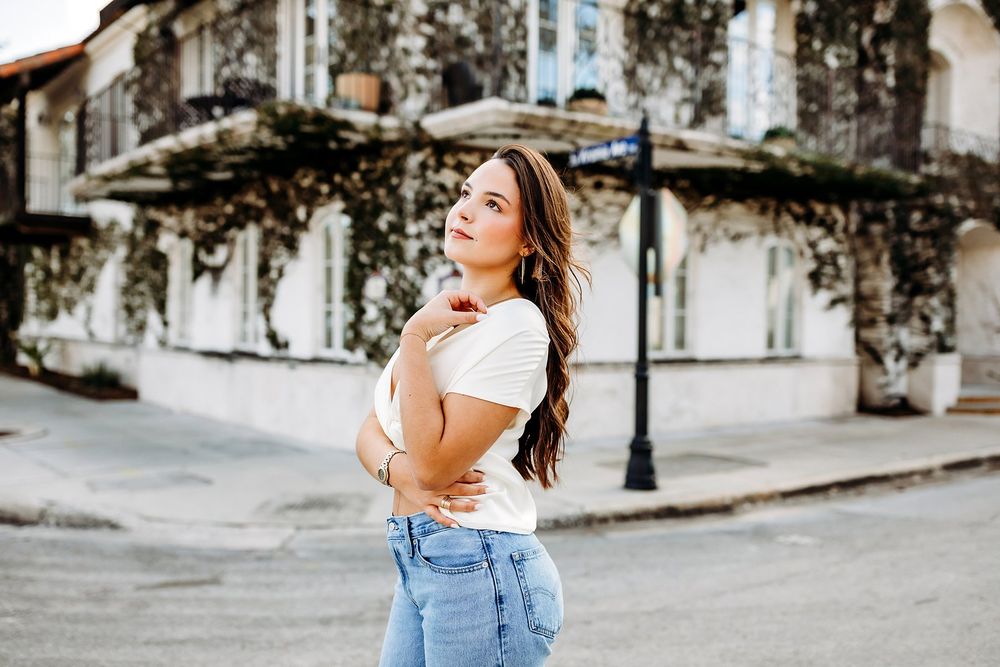 Young woman in white top and jeans poses on a charming street with ivy-covered building backdrop.