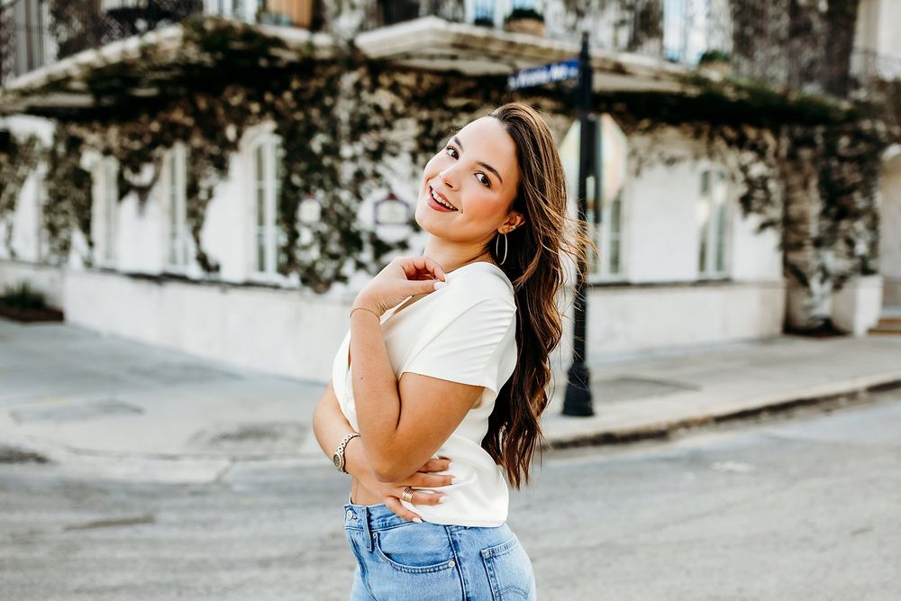 Young woman in white tee and jeans smiling over shoulder on a sunny street with ivy-covered building.