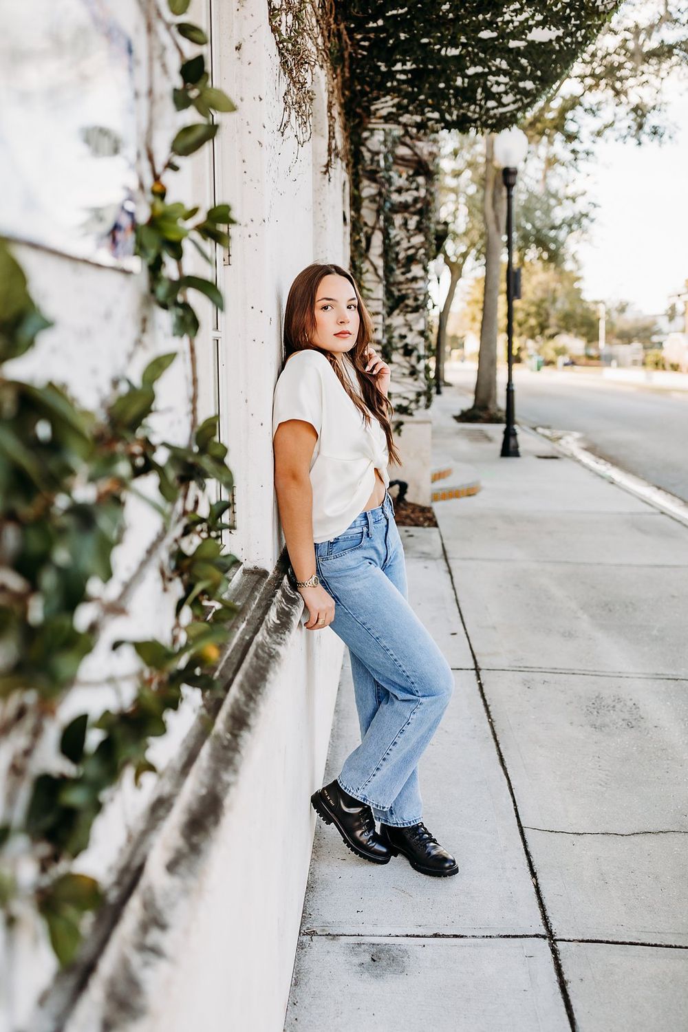 Young woman in white crop top and light jeans leaning against a white wall on a sidewalk surrounded by greenery.