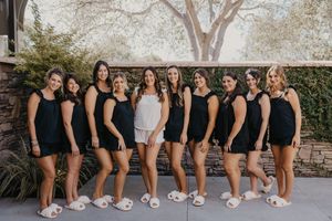Group of women in matching black rompers and white slippers posing together outdoors, with one woman in white center.