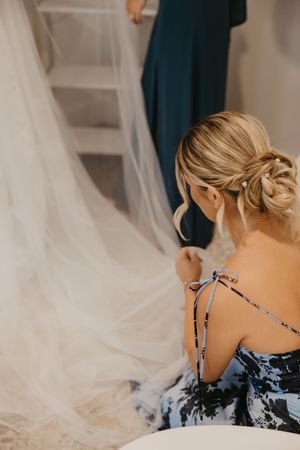 Blonde bridesmaid in floral blue dress arranging white wedding veil, elegant updo hairstyle with pearl pin.