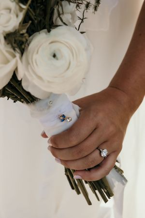 Bride holding white floral bouquet with diamond engagement ring and blue sapphire ring tied with ribbon.