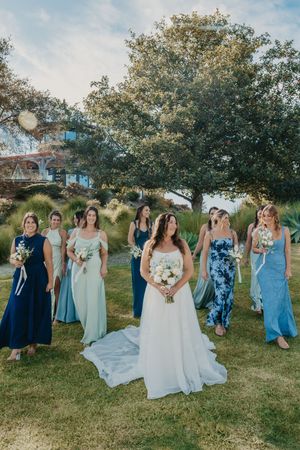 Bride in white gown walks with bridesmaids in blue dresses outdoors on green lawn near large tree.