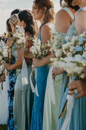 Bridesmaids in mismatched blue and green gowns holding white floral bouquets with ribbon streamers at an outdoor wedding.