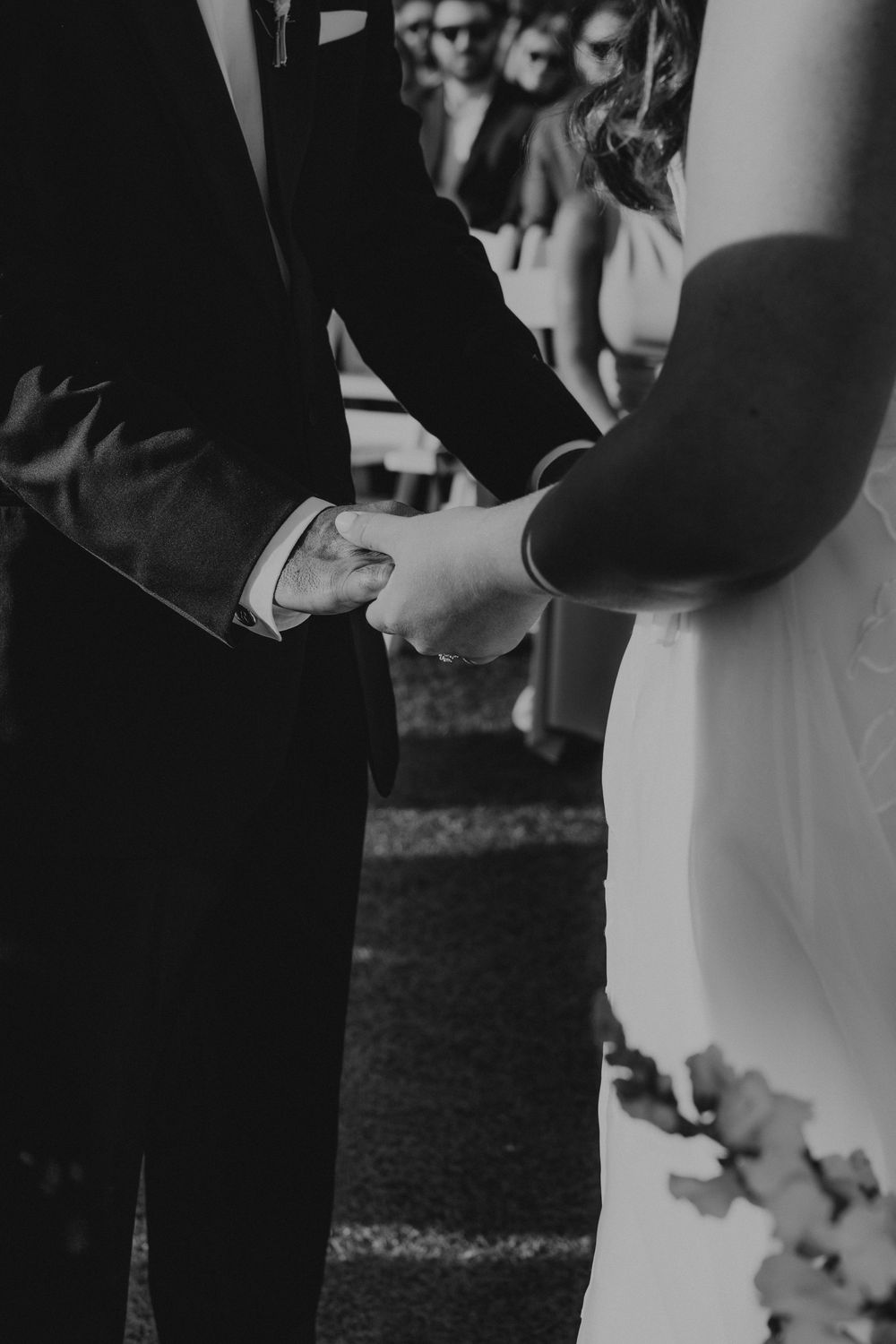 Black and white photo of a bride and groom holding hands during their wedding ceremony with guests in the background.