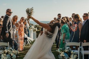 Bride celebrating in wedding aisle, raising bouquet joyfully, guests cheering outdoors near ocean backdrop.