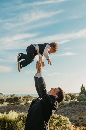 A father in a suit joyfully tosses his young son into the air outdoors under a blue sky.
