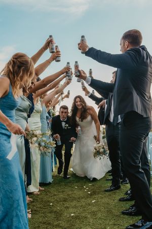 Bride and groom walk through tunnel of raised beer cans held by wedding party in blue dresses outdoors.