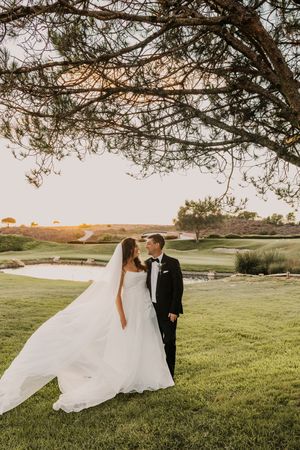 Bride and groom share a romantic moment under a tree at sunset on a lush golf course, veil flowing in the breeze.