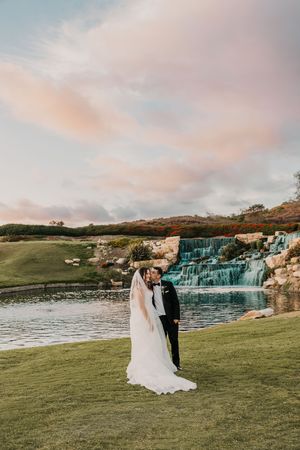Bride and groom share a romantic kiss at sunset near a waterfall and pond on a lush green golf course.