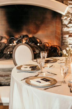 Elegant wedding table setting with gold chargers and white linens in front of a glowing stone fireplace.